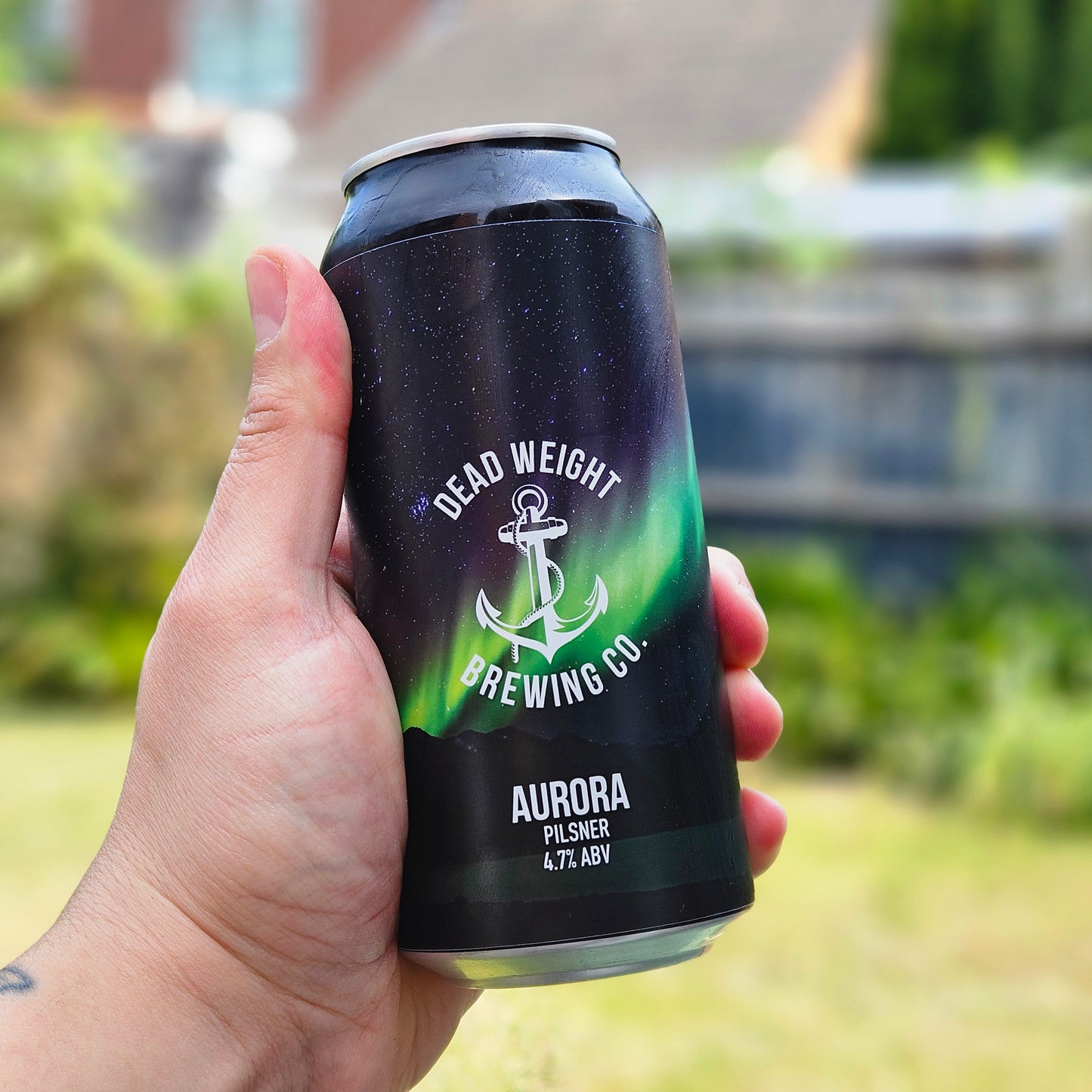 Hand holding a can of Aurora Pilsner from Dead Weight Brewing Co. beer with a blurred outdoor background